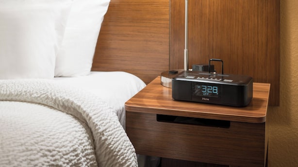 A neatly made bed with white linens, a wooden bedside table with a digital clock, and a lamp.
