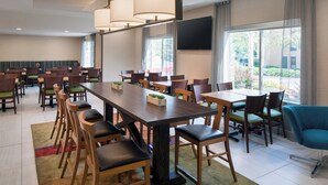 A modern hotel dining area with multiple tables and chairs, a large flat-screen TV on the wall, and large windows with curtains.