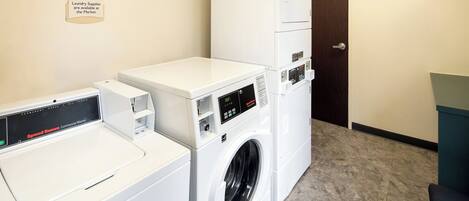 A laundry area with a Speed Queen commercial washer and dryer, a wall-mounted sign indicating cycle times and laundry supplies availability, and a door leading to another room.