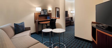 A hotel room with a sofa, a desk with a chair, two small tables, a television, and a patterned carpet.
