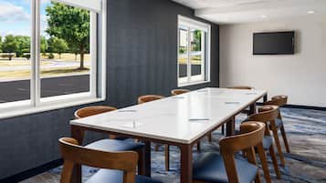 Une salle de conférence avec une longue table, des chaises, un téléviseur fixé au mur et de grandes fenêtres donnant sur des arbres et un parking.