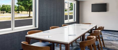 A conference room with a long table, chairs, a TV on the wall, and large windows with a view of trees and a parking lot.