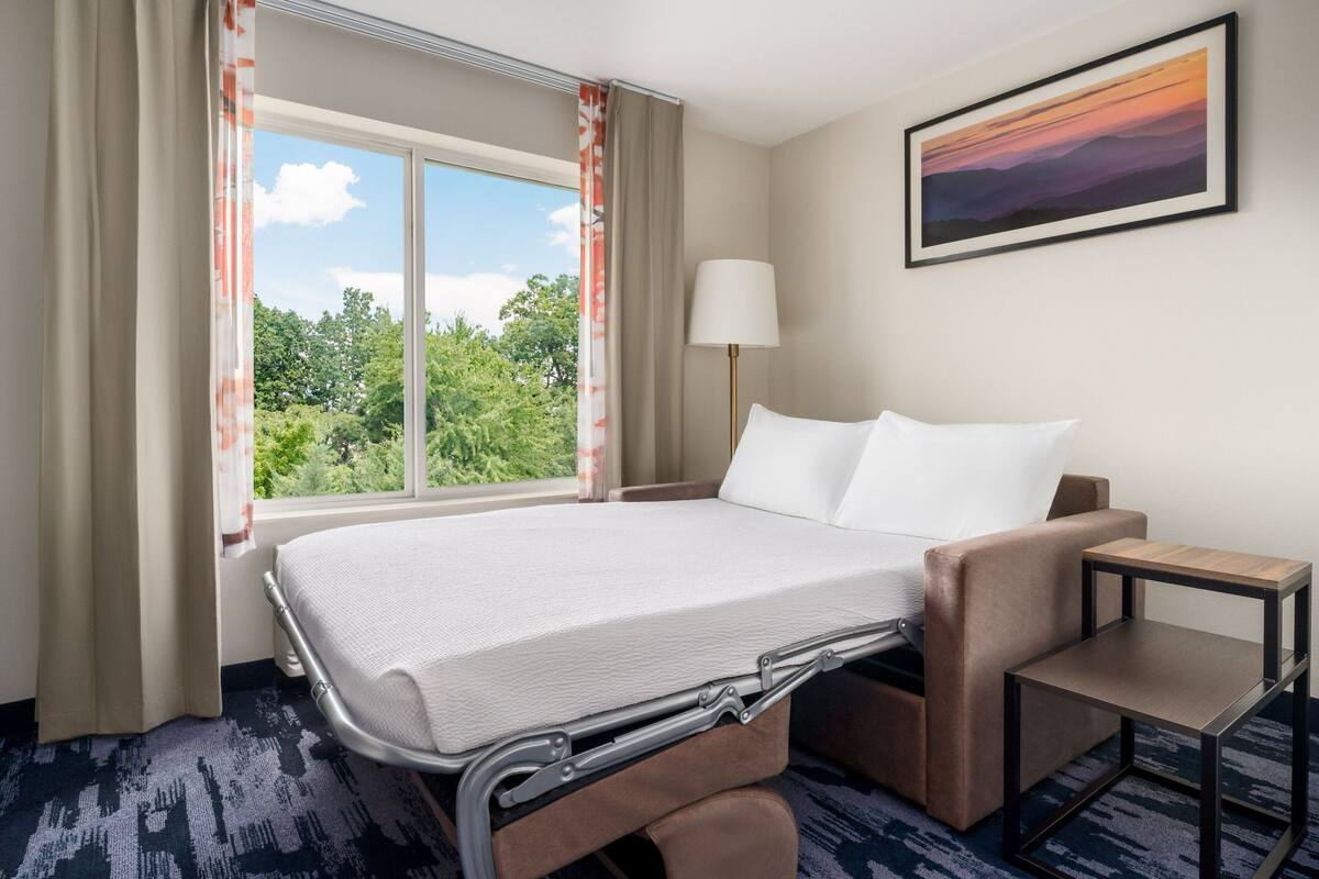 Une chambre d’hôtel avec un lit, un bureau, une chaise, une lampe et une fenêtre avec des rideaux.