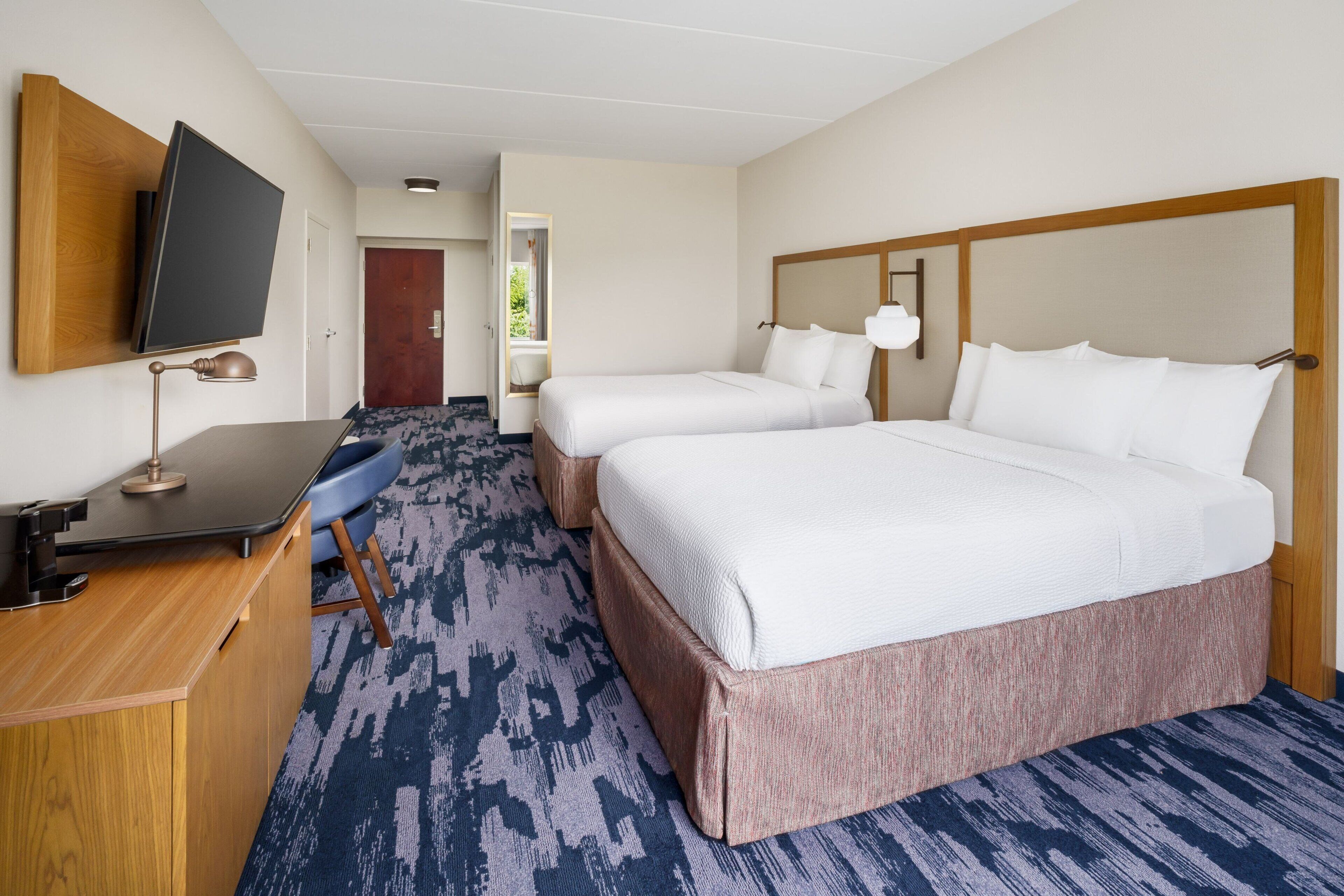 A hotel room with two beds, a desk, a TV mounted on the wall, and a patterned carpet.