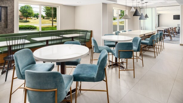 Un comedor moderno con mesas redondas y sillas azules, una zona de asientos tipo banco en tonos verdes y amplios ventanales con vistas al exterior.