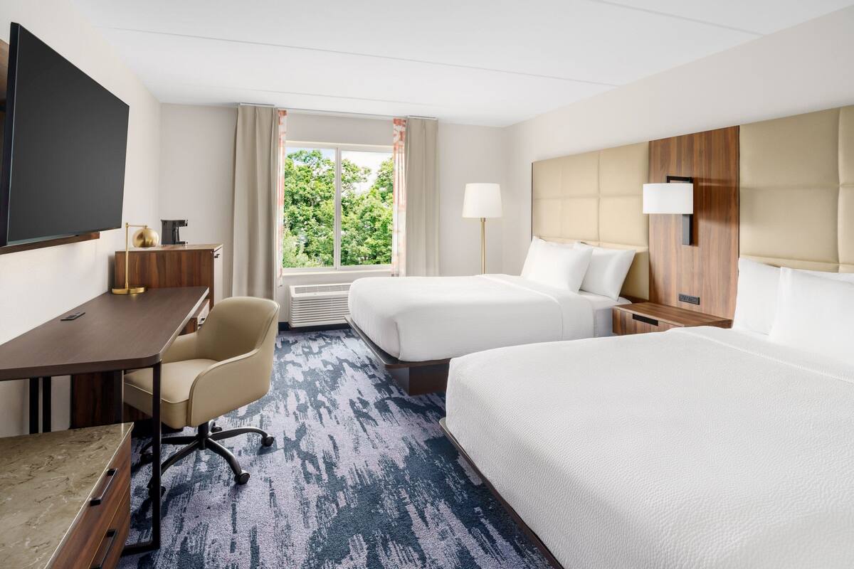Une chambre d’hôtel équipée d’un bureau, de deux lits, d’une télévision, d’une fenêtre avec des rideaux et d’une lampe.