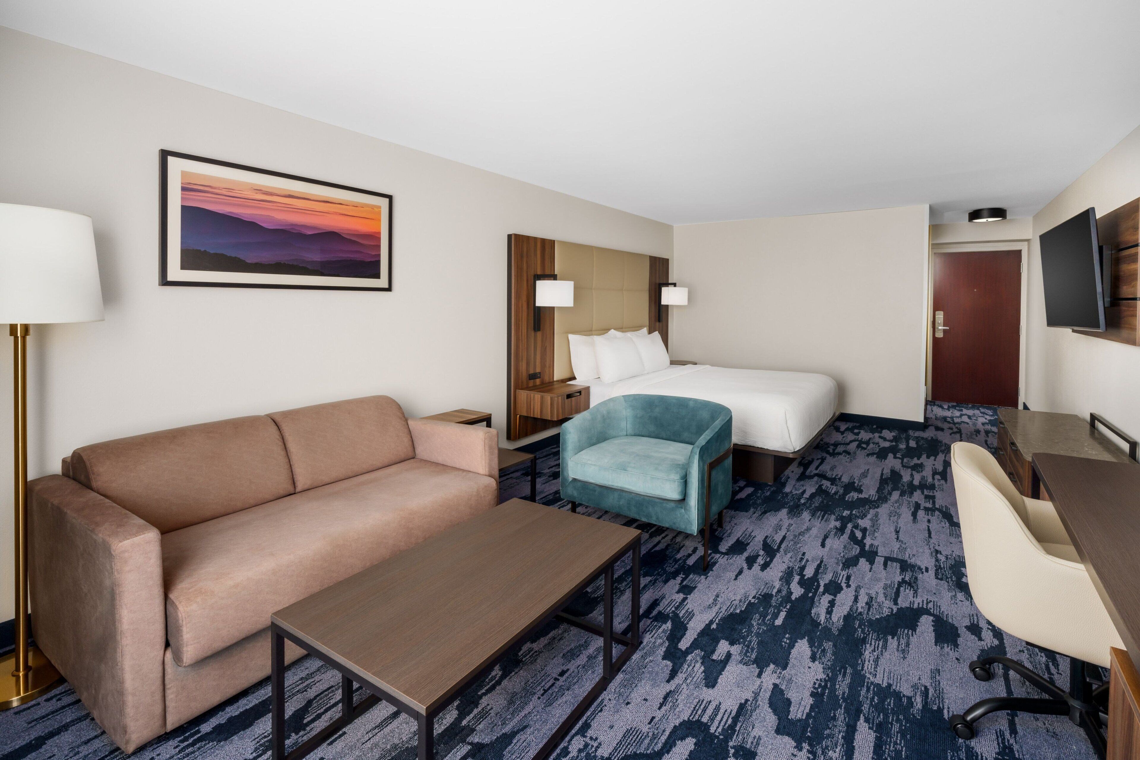 A hotel room with a sofa, a bed, a desk, and a chair. There is a painting on the wall and a TV mounted on the wall.