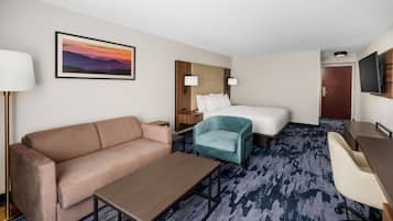 A hotel room with a sofa, a bed, a desk, and a chair. There is a painting on the wall and a TV mounted on the wall.