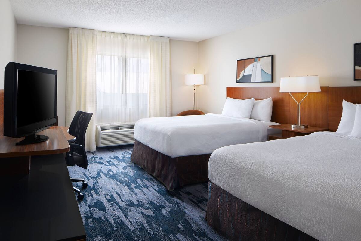 A hotel room with two beds, a desk with a television, and a large window with curtains.