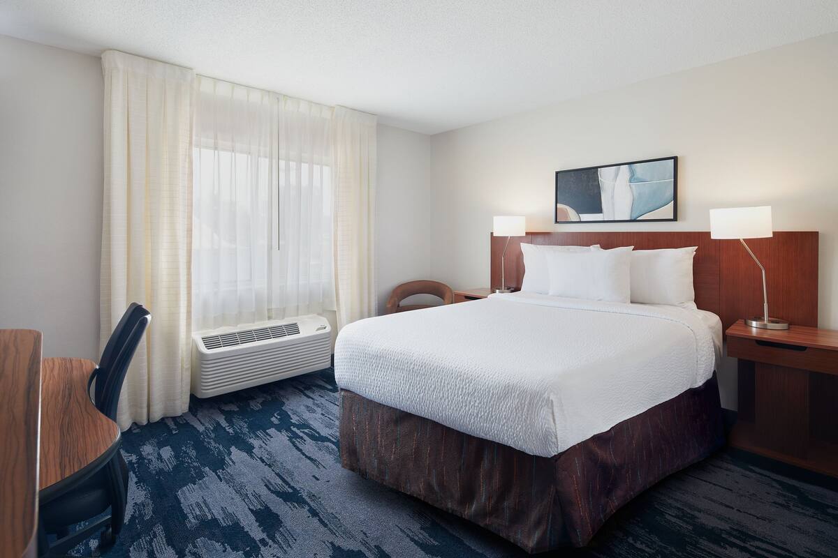 A hotel room with a large bed, a desk, a chair, a window with sheer curtains, a wall-mounted air conditioner, and a painting above the bed.