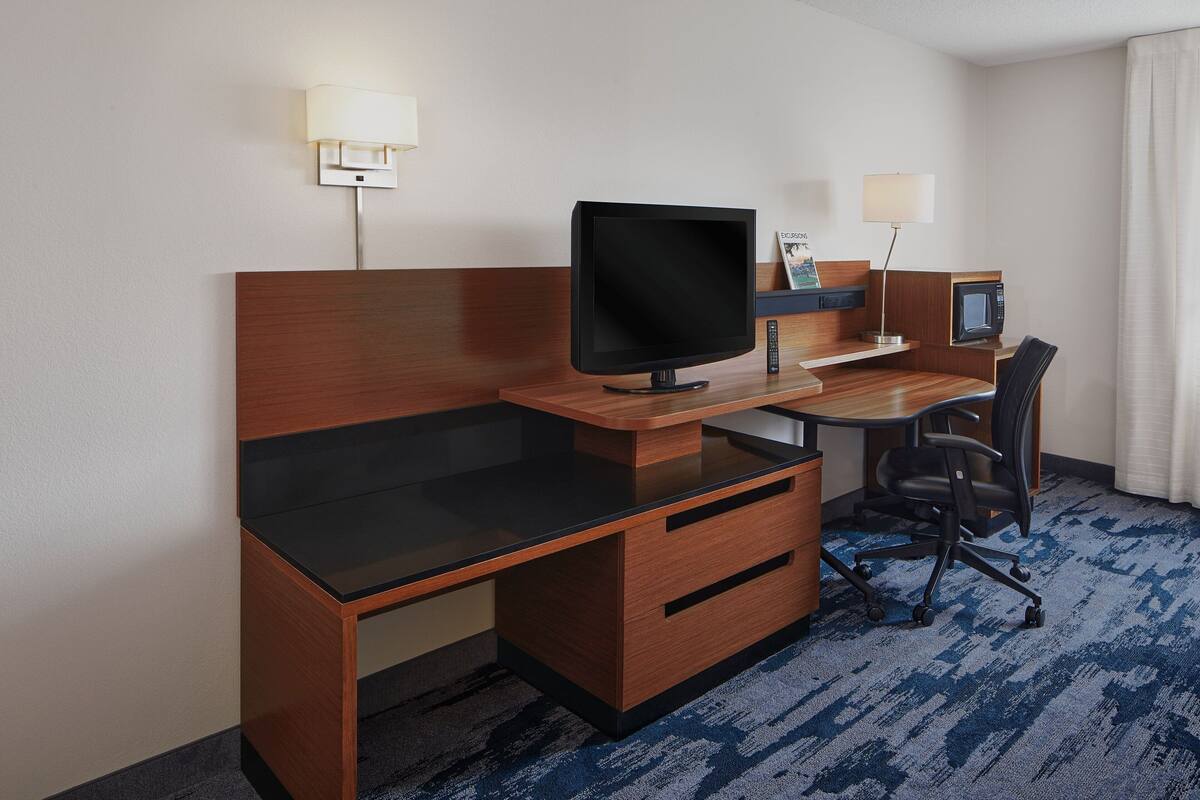 A hotel room with a wooden desk, a flat-screen TV, a microwave, a chair, and a lamp.
