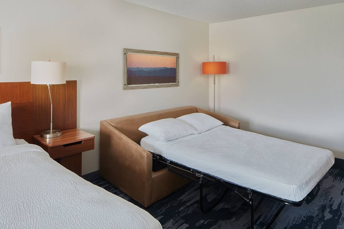 A hotel room with a single bed, a nightstand with a lamp, a wall-mounted painting, and a separate reading light.