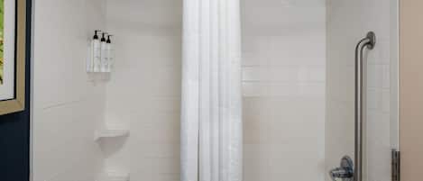 A shower area with a white shower curtain, a handheld showerhead, and a grab bar on the right. There are two white shelves on the left wall, and a framed picture above them.