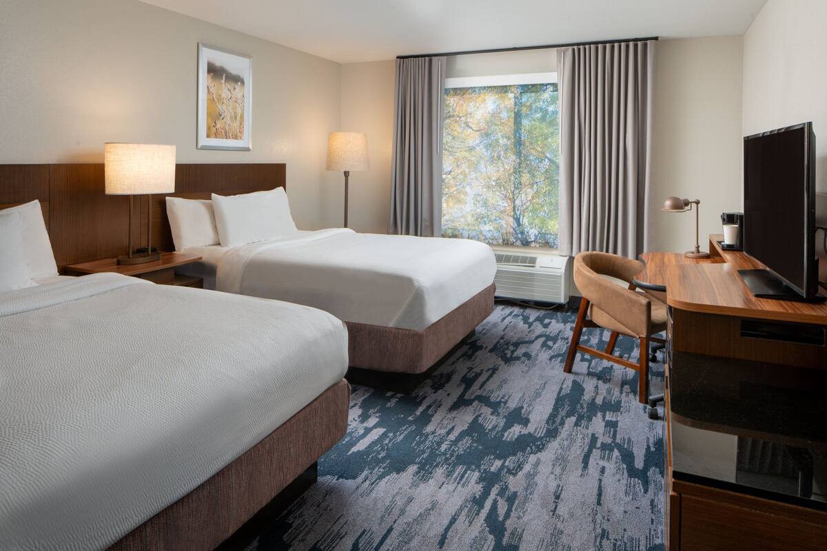 A hotel room with two beds, a desk, a television, and a window with curtains.