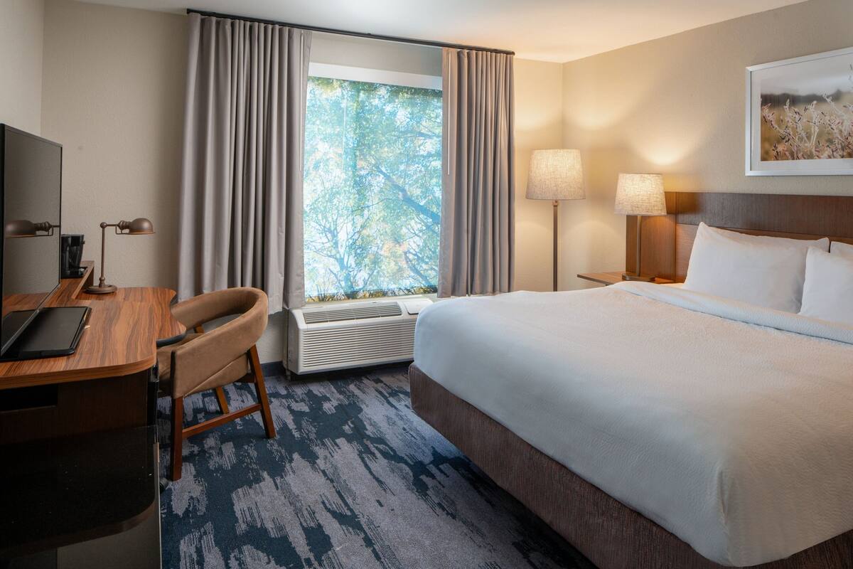 A hotel room with a bed, desk, chair, air conditioning unit, and a window with curtains.