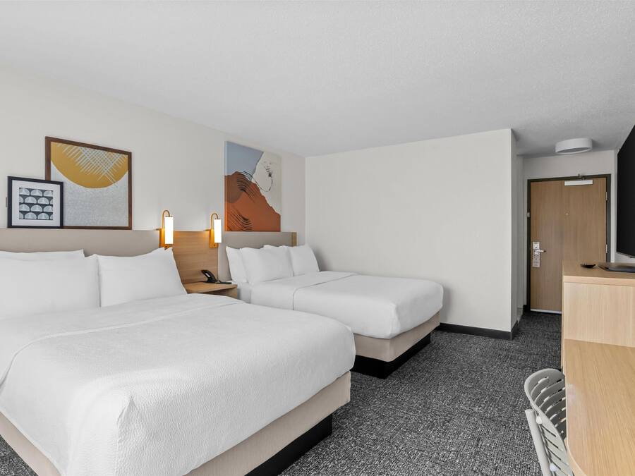 Habitación de hotel con dos camas, un escritorio, una silla, un televisor y obras de arte en las paredes.