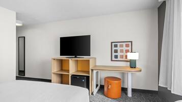 A hotel room with a bed, a TV on a wooden stand, a desk with a lamp, a small orange stool, and a framed picture on the wall.