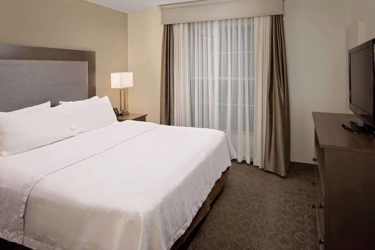 Une chambre d’hôtel avec un grand lit, une table de chevet avec une lampe, un téléviseur posé sur une commode et une fenêtre avec des rideaux.
