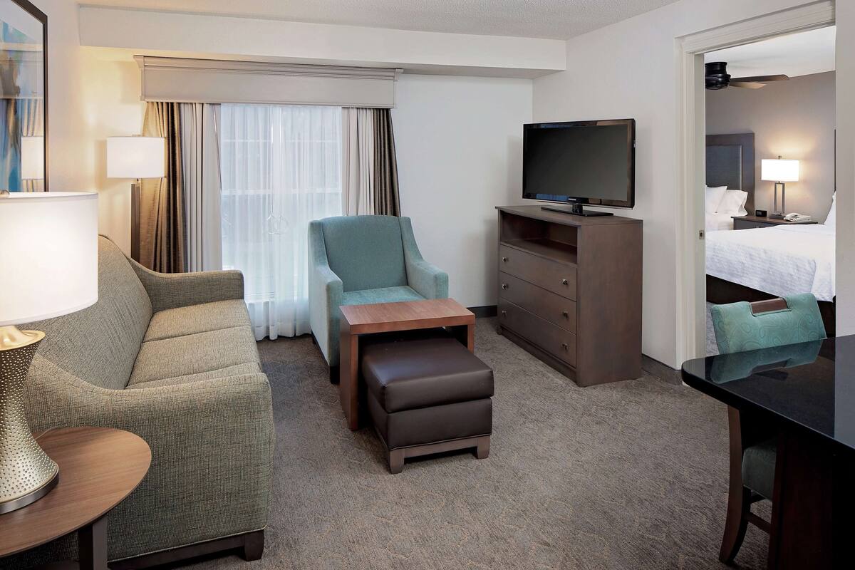 Une chambre d’hôtel comprenant un canapé, un fauteuil, un pouf, une télévision, une commode et un lit avec des oreillers et une table de chevet.