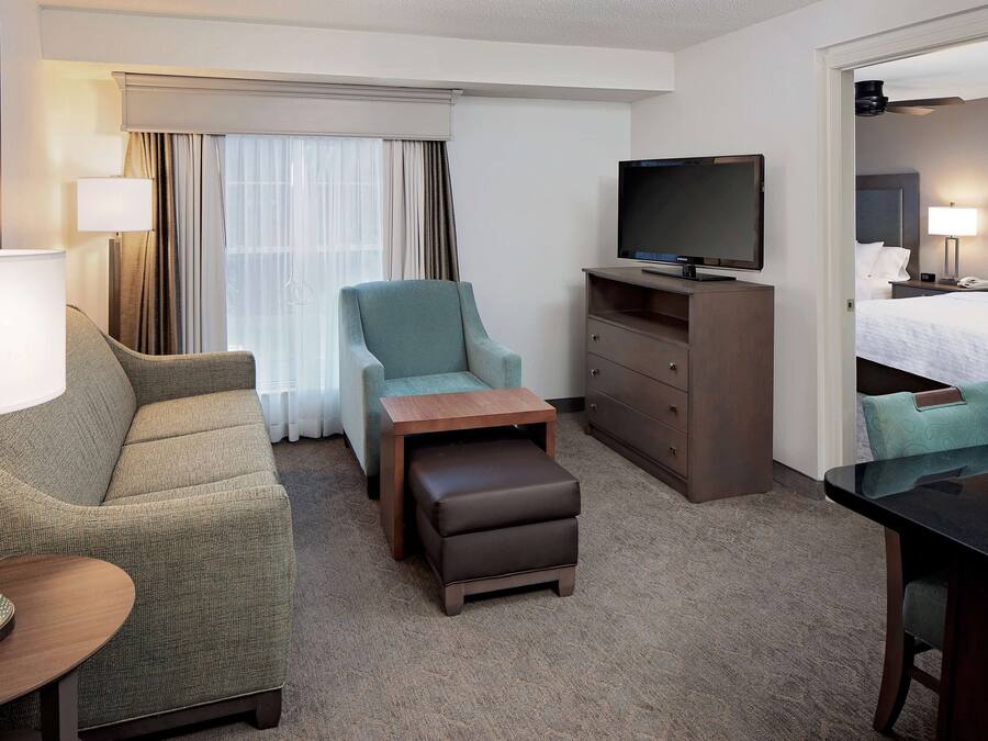 A hotel room with a sofa, armchair, ottoman, TV, dresser, and a bed with pillows and a nightstand.
