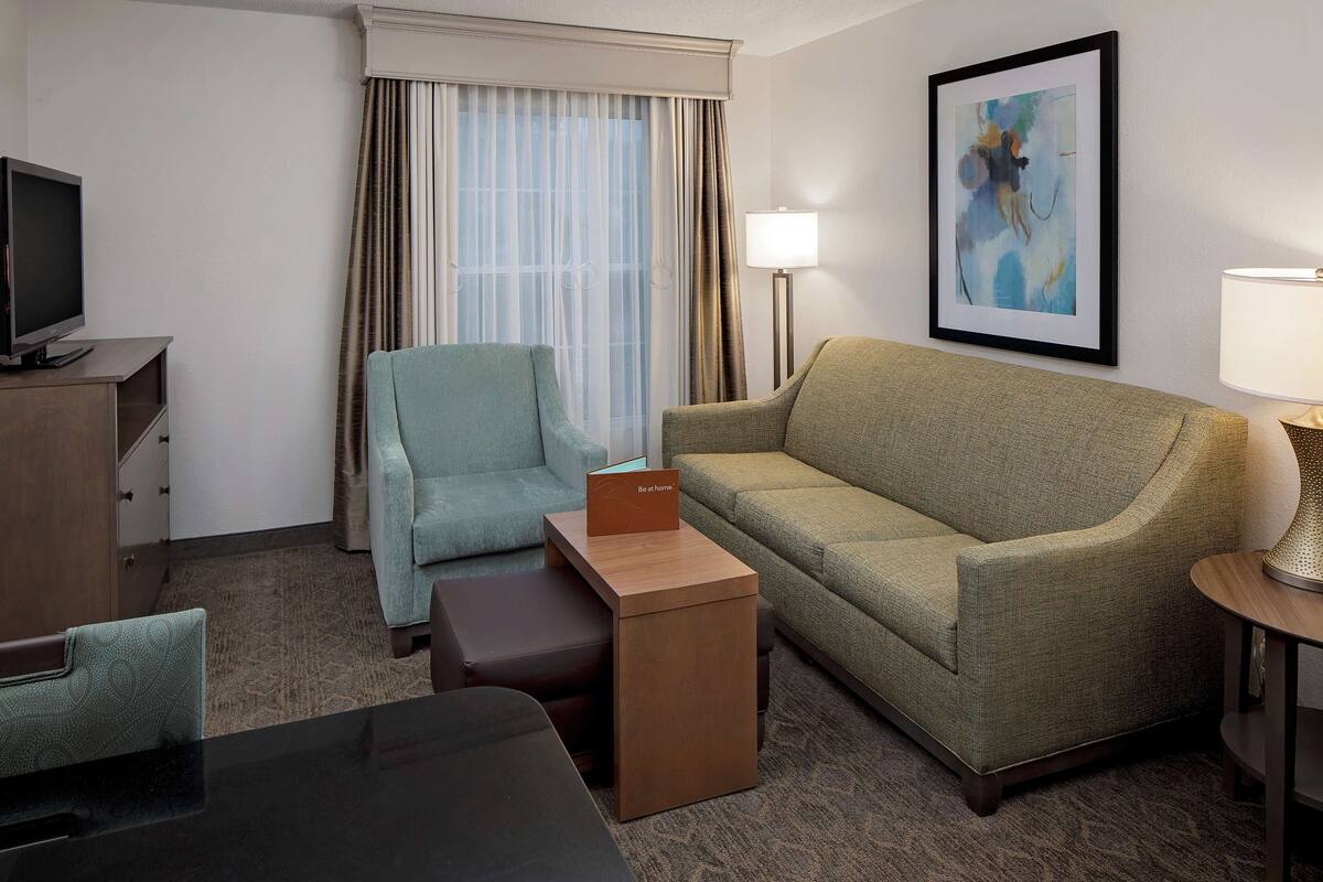 Une chambre d’hôtel avec un canapé, un fauteuil, une petite table, un téléviseur et un tableau au mur.
