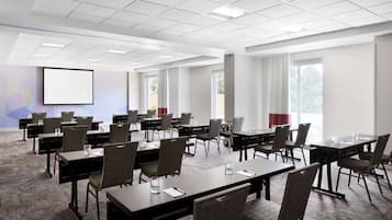 Une salle de conférence équipée de plusieurs tables et chaises, d’un écran de projection et de grandes fenêtres.