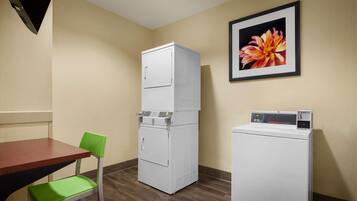 A hotel room with a wooden table, a green chair, a stacked washer and dryer, and a framed picture on the wall.