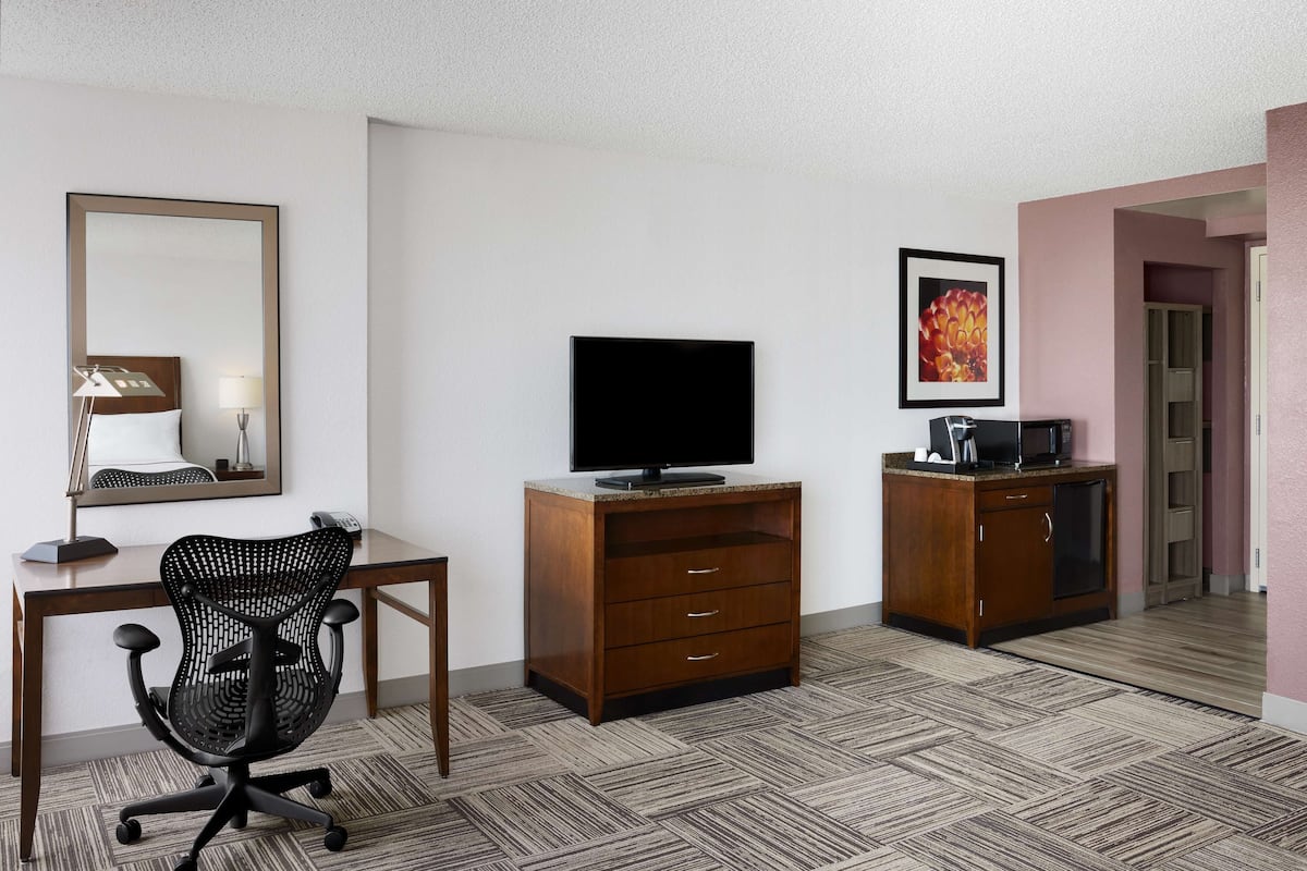 Een hotelkamer met een houten bureau, een zwarte bureaustoel, een televisie op een houten kast en een kleine kitchenette.