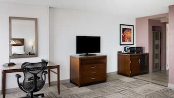 A hotel room with a wooden desk, a black office chair, a TV on a wooden cabinet, and a small kitchenette.