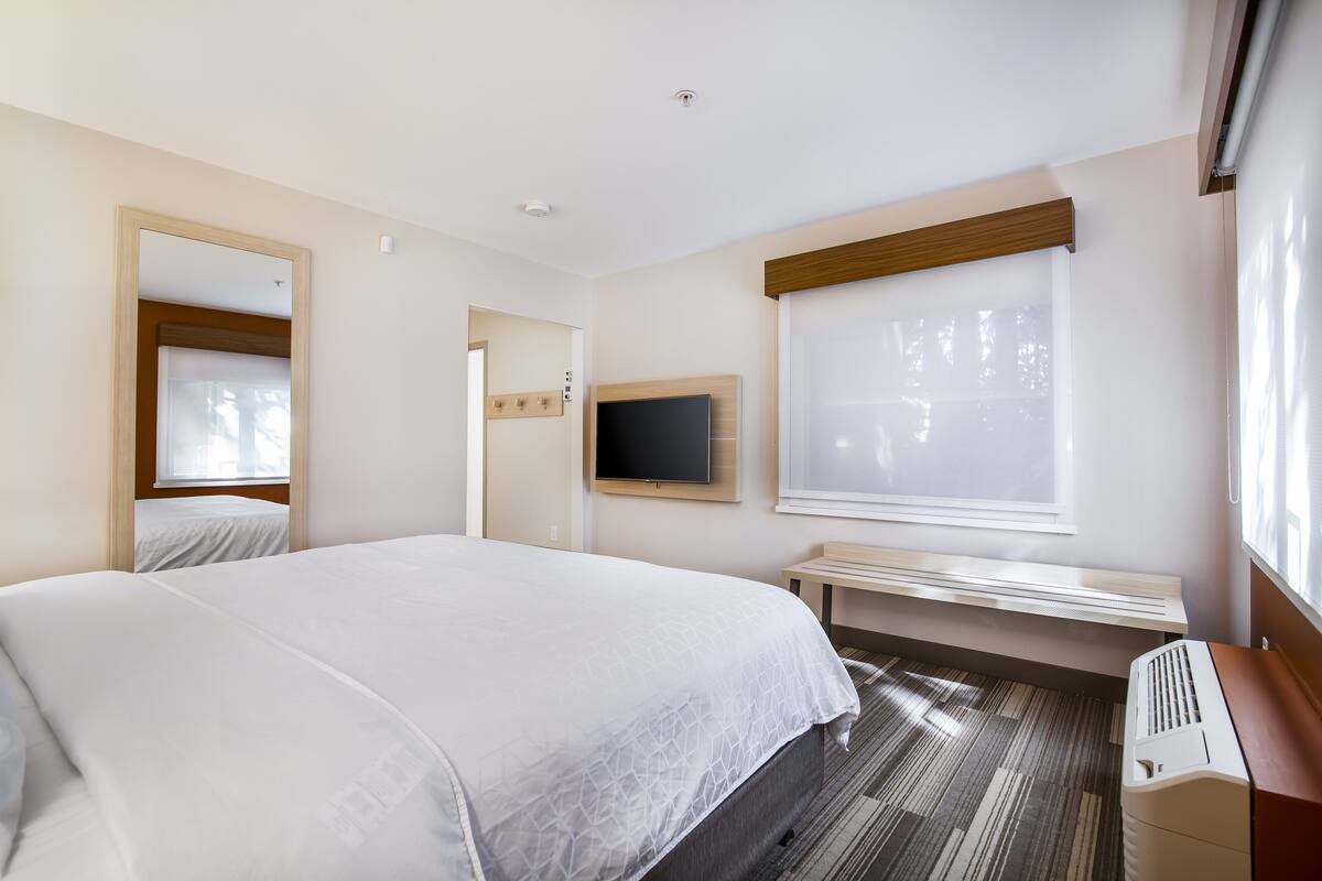 Une chambre d’hôtel avec un grand lit, un téléviseur fixé au mur, un banc et une fenêtre avec des stores.