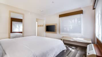 A hotel room with a large bed, a TV mounted on the wall, a bench, and a window with blinds.
