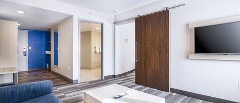 A modern hotel room with a sofa, a coffee table, a TV mounted on the wall, and a sliding door leading to a bathroom.