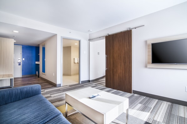 A modern hotel room with a sofa, a coffee table, a TV mounted on the wall, and a sliding door leading to a bathroom.