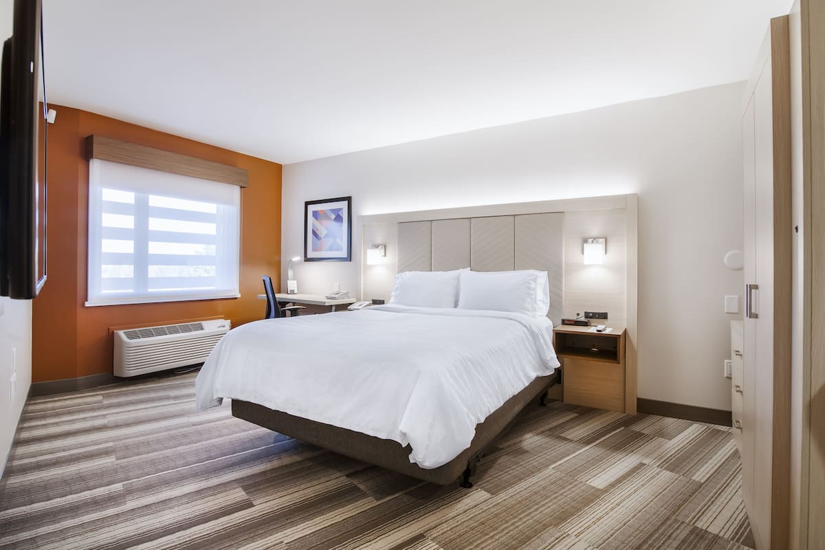 Une chambre d’hôtel avec un grand lit, une table de chevet, une commode, une fenêtre avec des stores, une lampe fixée au mur et un tableau encadré.