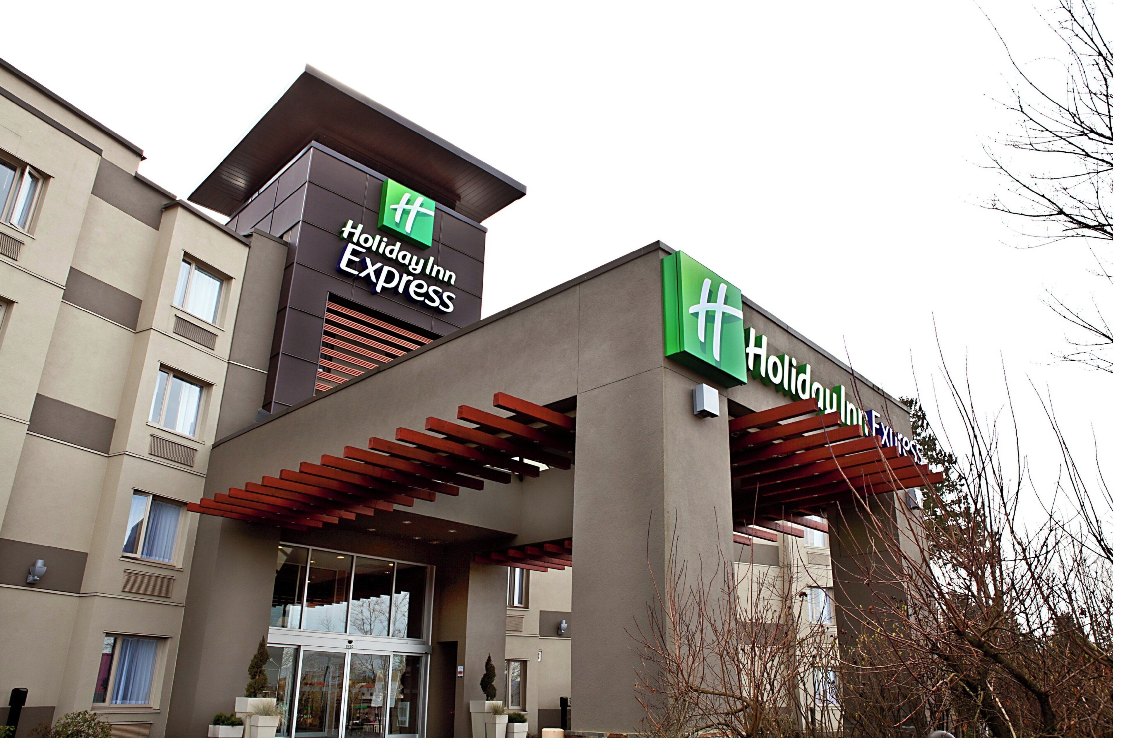 A Holiday Inn Express building with a modern design, including a prominent green and white sign, large windows, and a covered entrance area.
