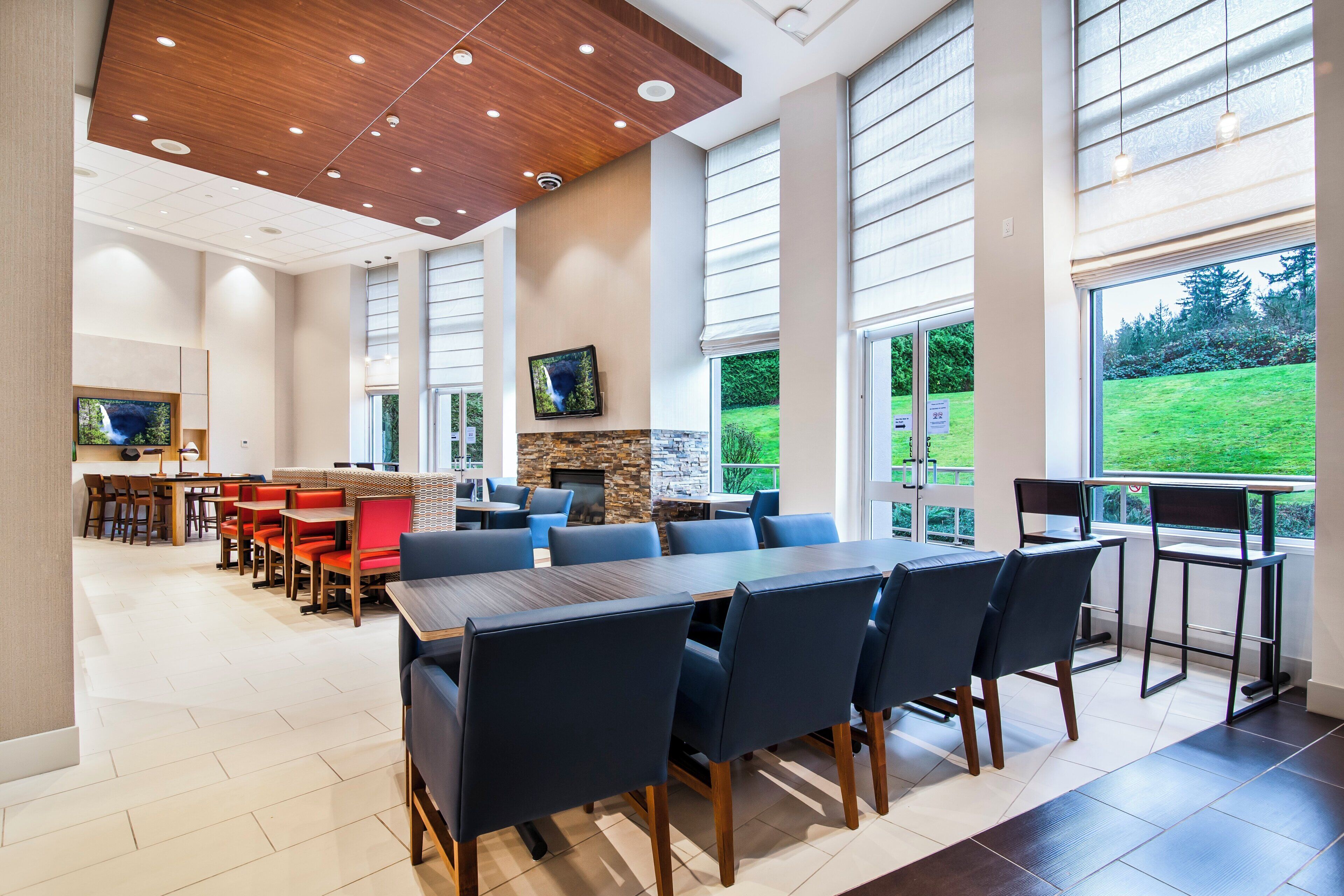 A spacious indoor area with a long table and chairs, a television mounted on the wall, and large windows providing a view of the outdoors.