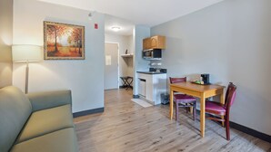 A modern living room with a sofa, a dining table with chairs, a kitchen area with cabinets and a microwave, and a painting on the wall.