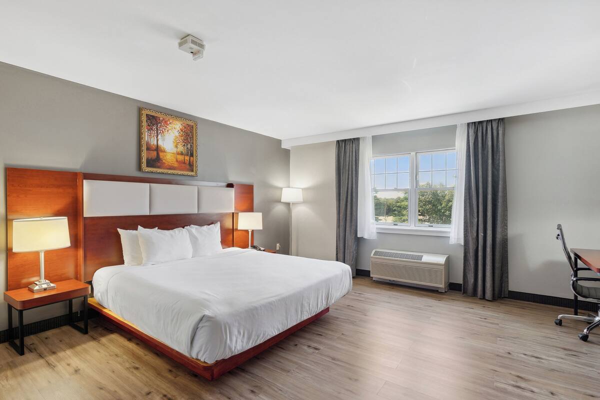 A hotel room with a large bed, two bedside tables with lamps, a window with curtains, and a painting on the wall.