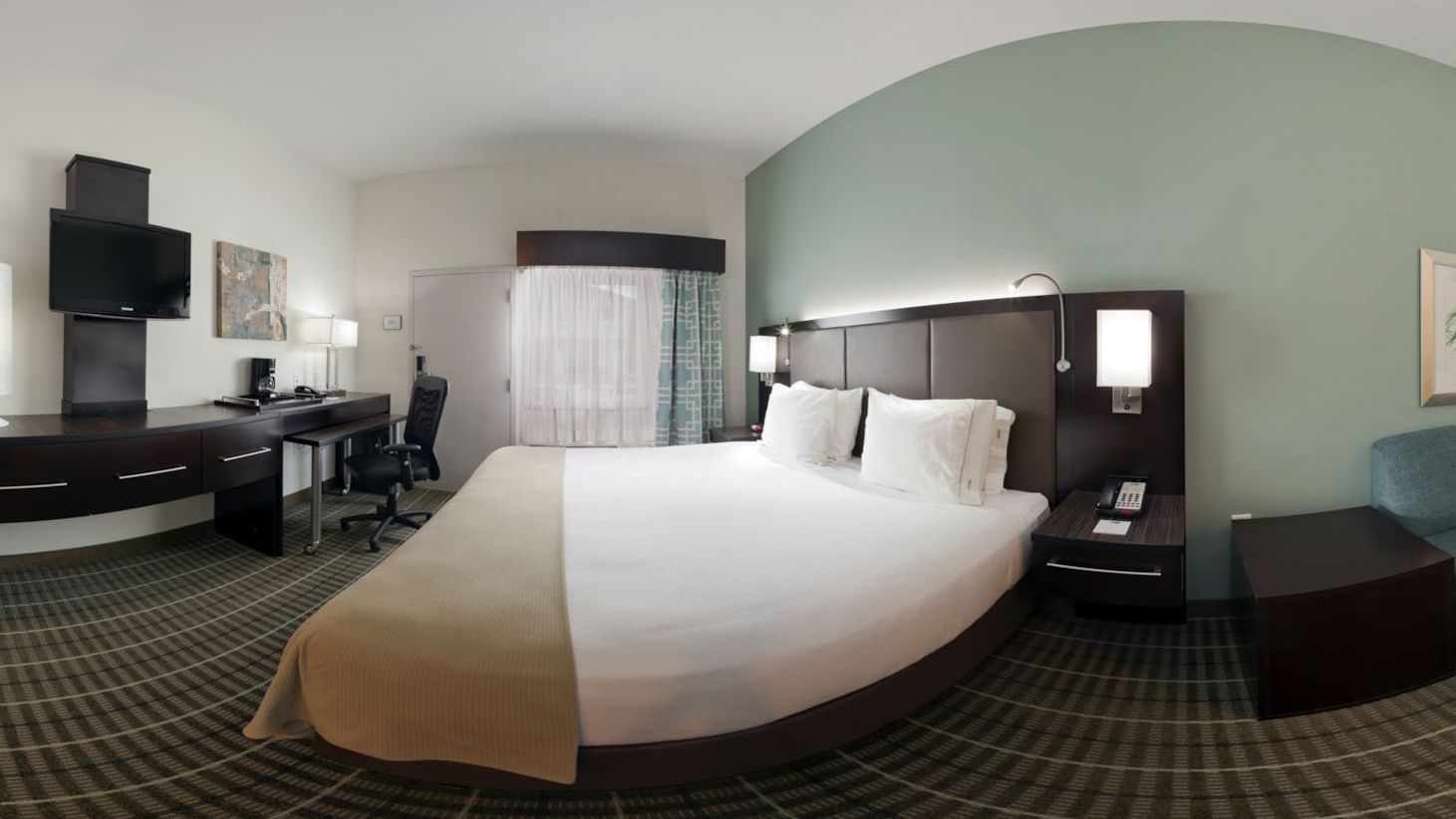 A hotel room with a large bed, a desk with a chair, a TV, and a nightstand with a lamp.