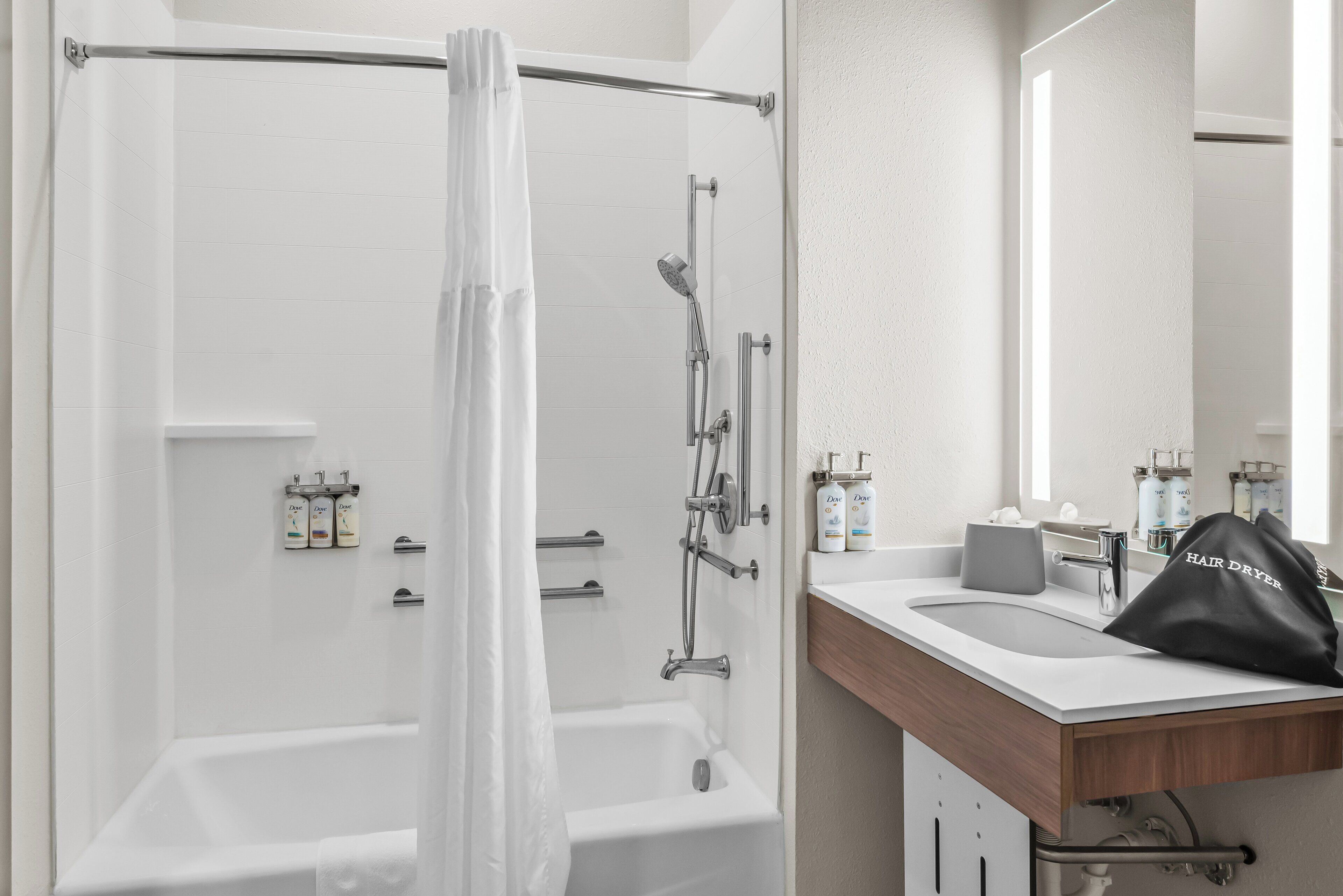 A modern bathroom with a bathtub, shower, and sink. There are toiletries on the counter and a hair dryer bag.
