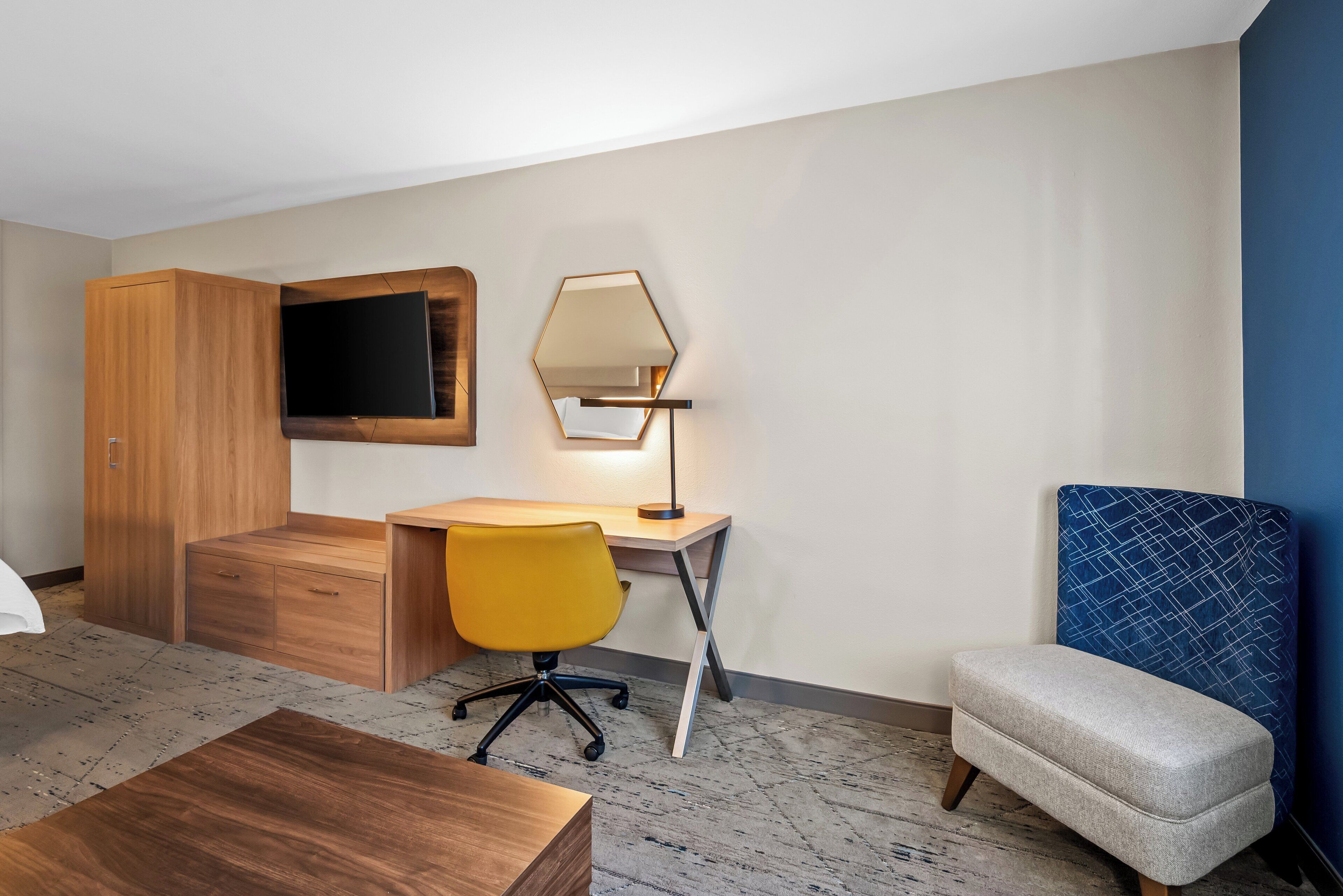A hotel room with a wooden wardrobe, a desk with a yellow chair, a blue upholstered chair, a flat-screen TV, and a floor lamp.