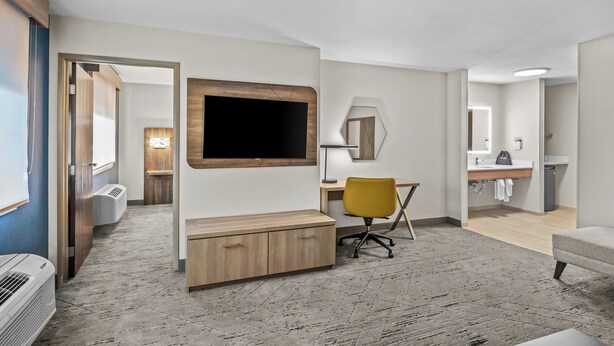 A modern hotel room with a large flat-screen TV mounted on the wall, a wooden desk with a chair, a small seating area with a sofa, and a bathroom visible through an open door. - Holiday Inn Express Monticello by IHG (Monticello)