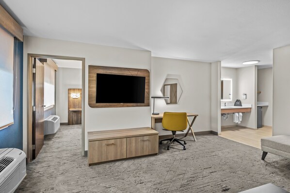 A modern hotel room with a large flat-screen TV mounted on the wall, a wooden desk with a chair, a small seating area with a sofa, and a bathroom visible through an open door. - Holiday Inn Express Monticello by IHG (Monticello)