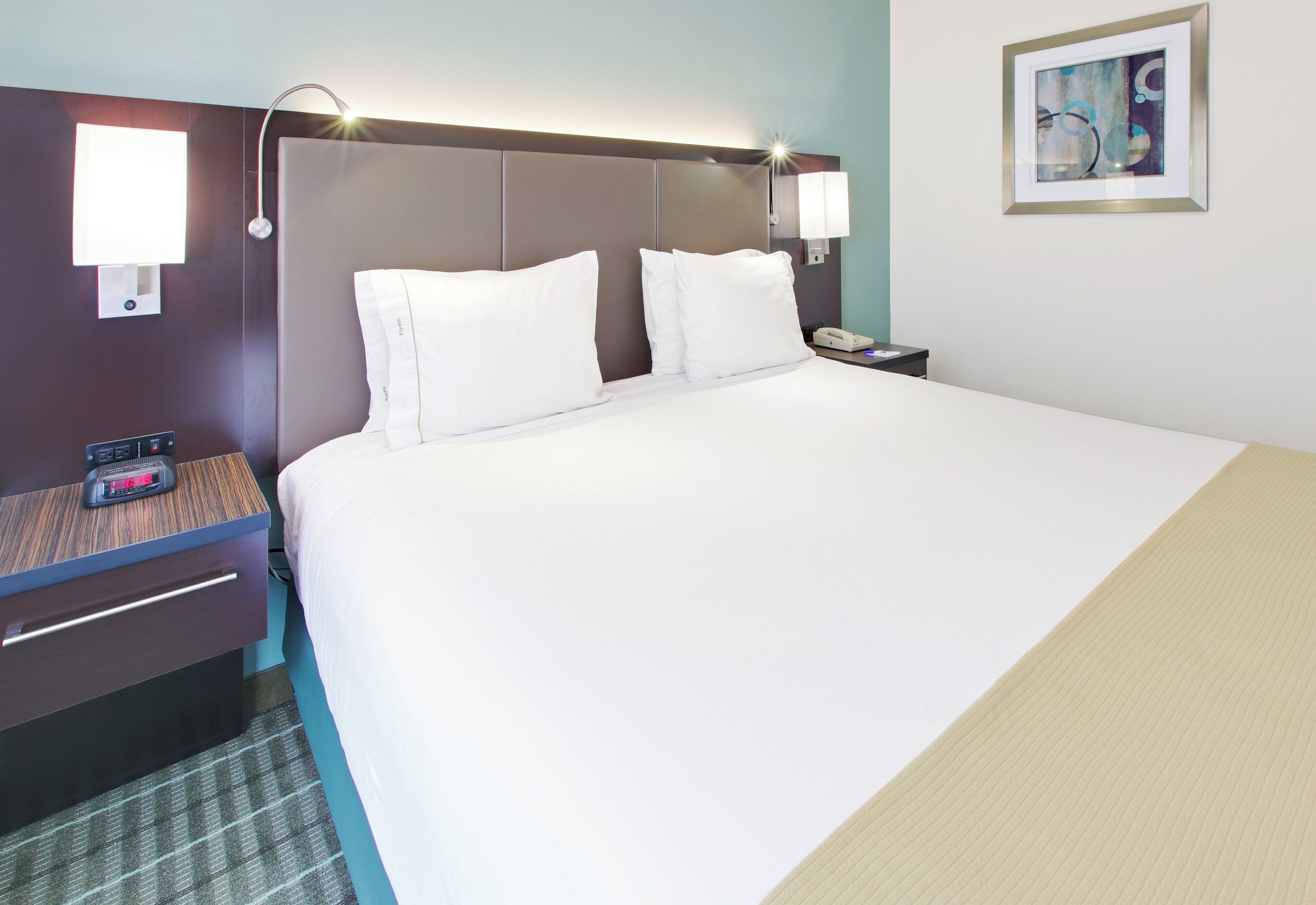 A hotel room with a large bed, two bedside tables, a nightstand, a lamp, a clock, and a painting on the wall.