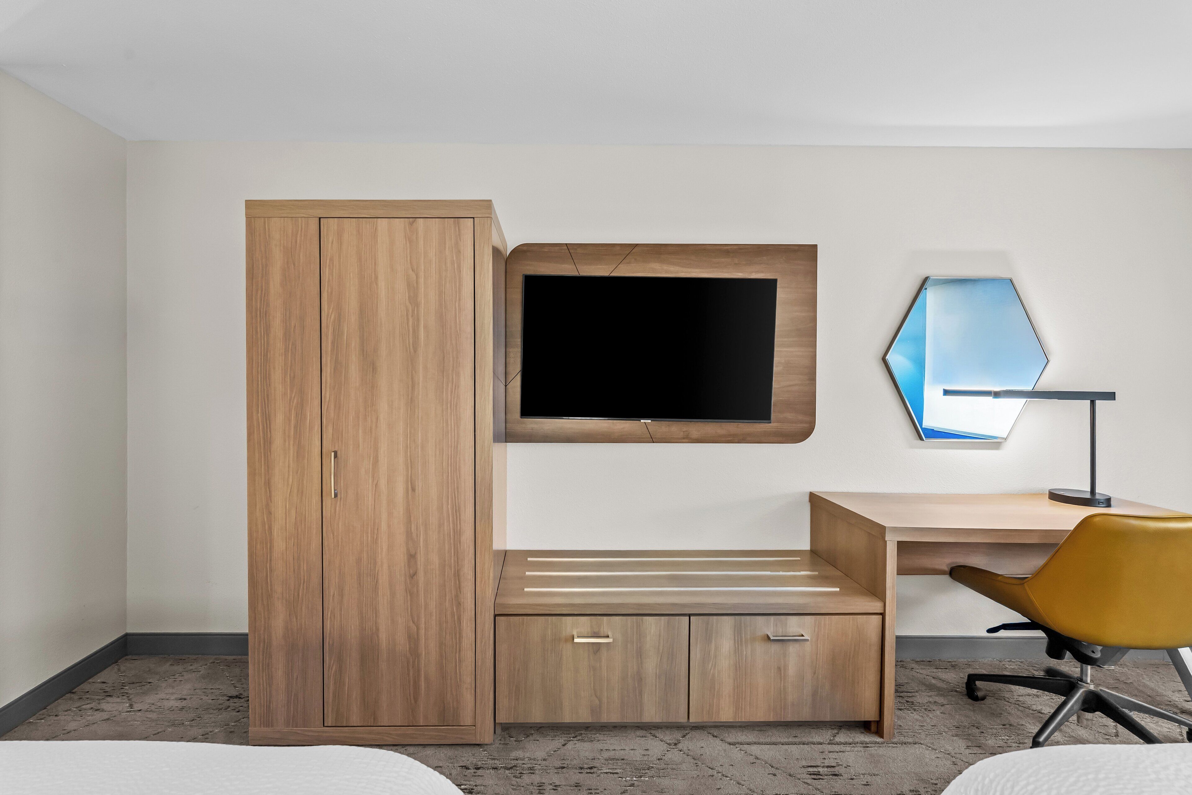 Un dormitorio con un armario de madera, un escritorio con silla, un televisor de pantalla plana y un espejo montado en la pared.
