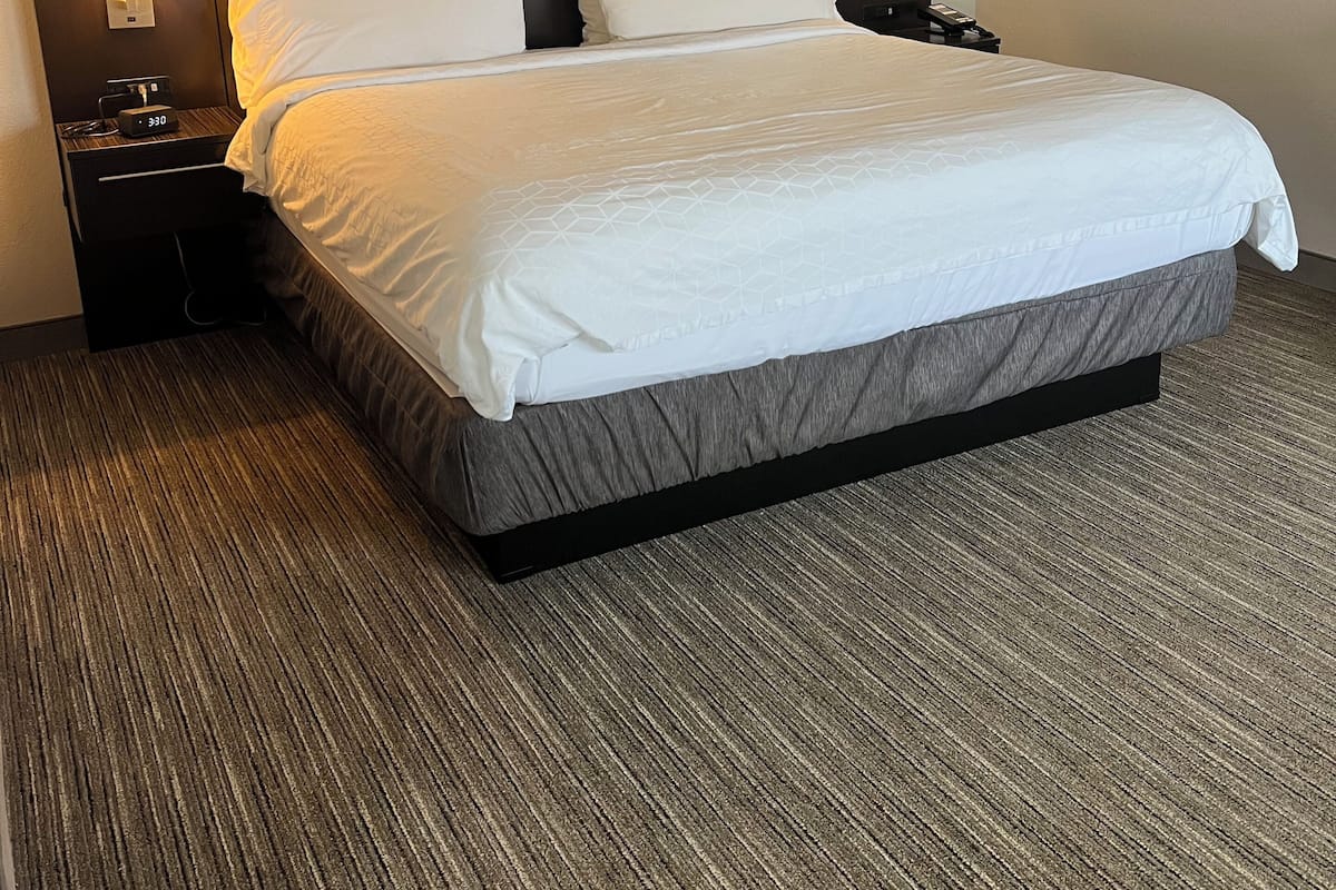 A hotel room with a large bed, two bedside tables with lamps, a wall-mounted alarm clock, and a headboard with integrated lighting.