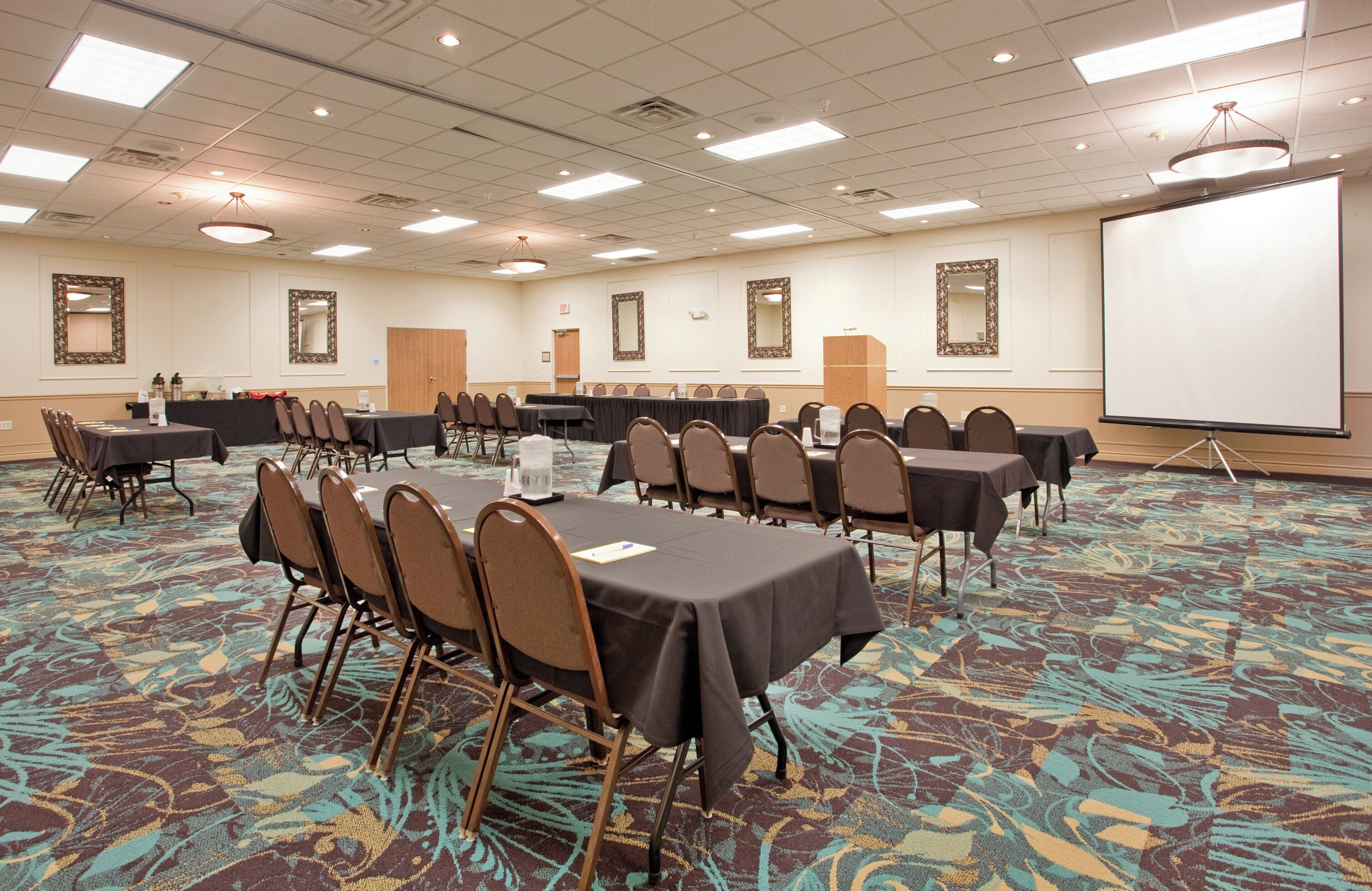 Une salle de conférence dotée de plusieurs tables et chaises, d’un écran de projection et d’un sol recouvert de moquette.