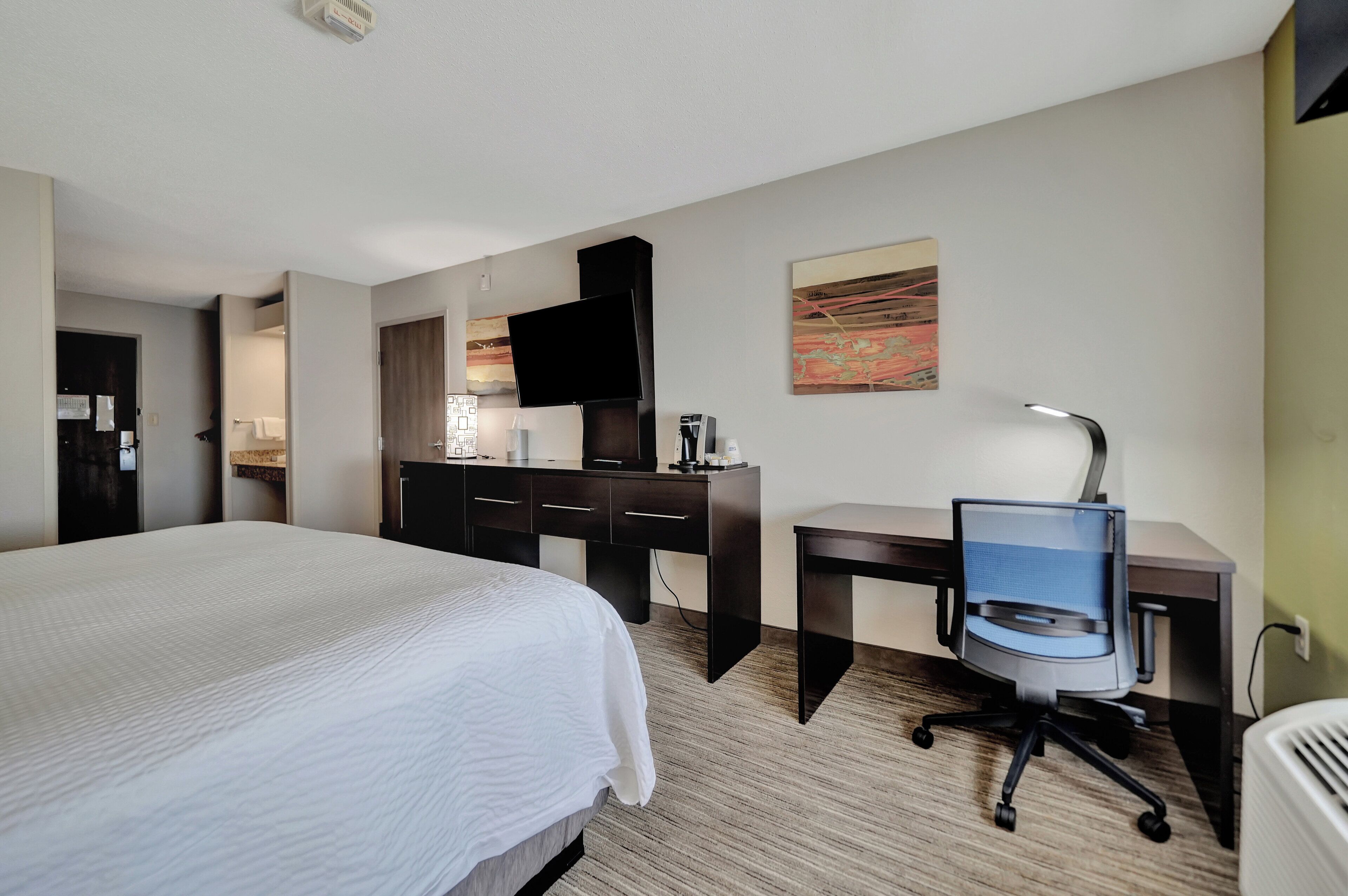 A hotel room with a large bed, a desk with a chair, a flat-screen TV, and a coffee machine.