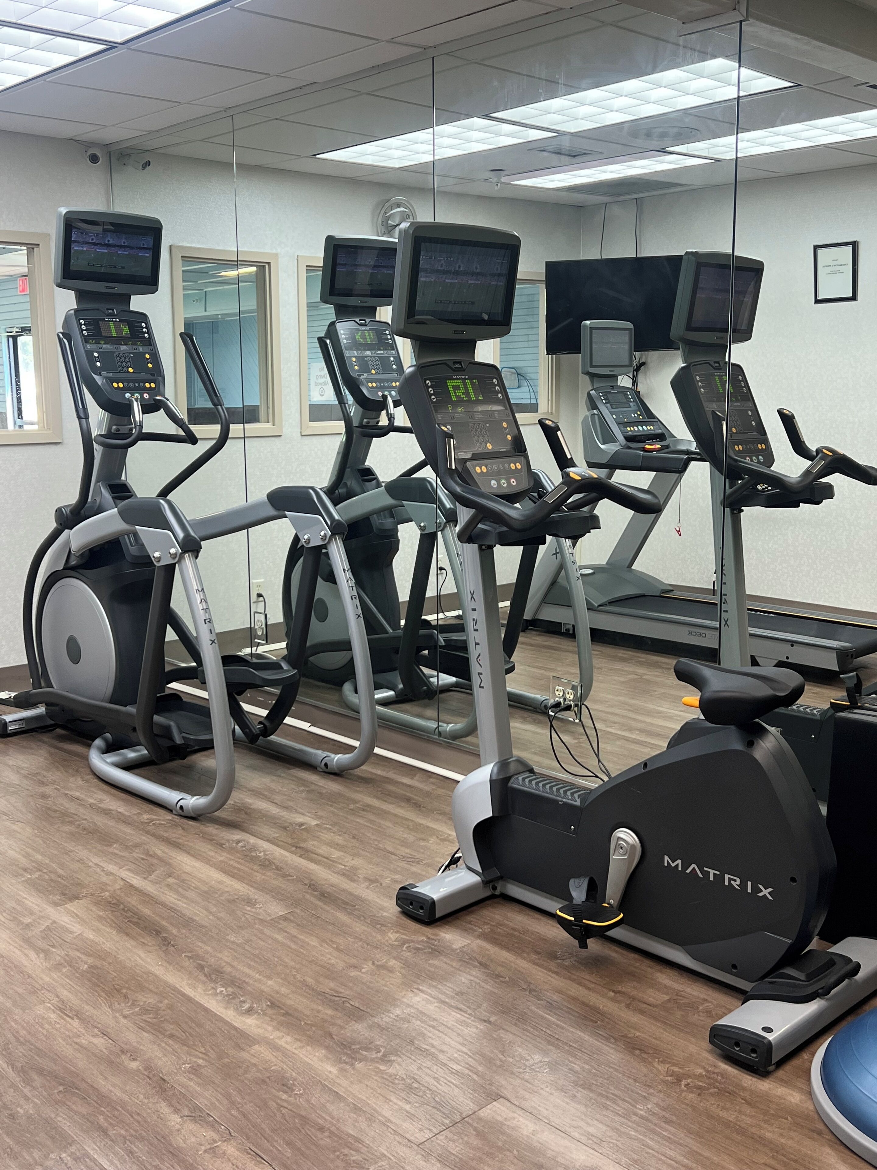 A row of stationary bikes with digital displays, a treadmill, and a rowing machine in a gym with wooden flooring and a white ceiling.