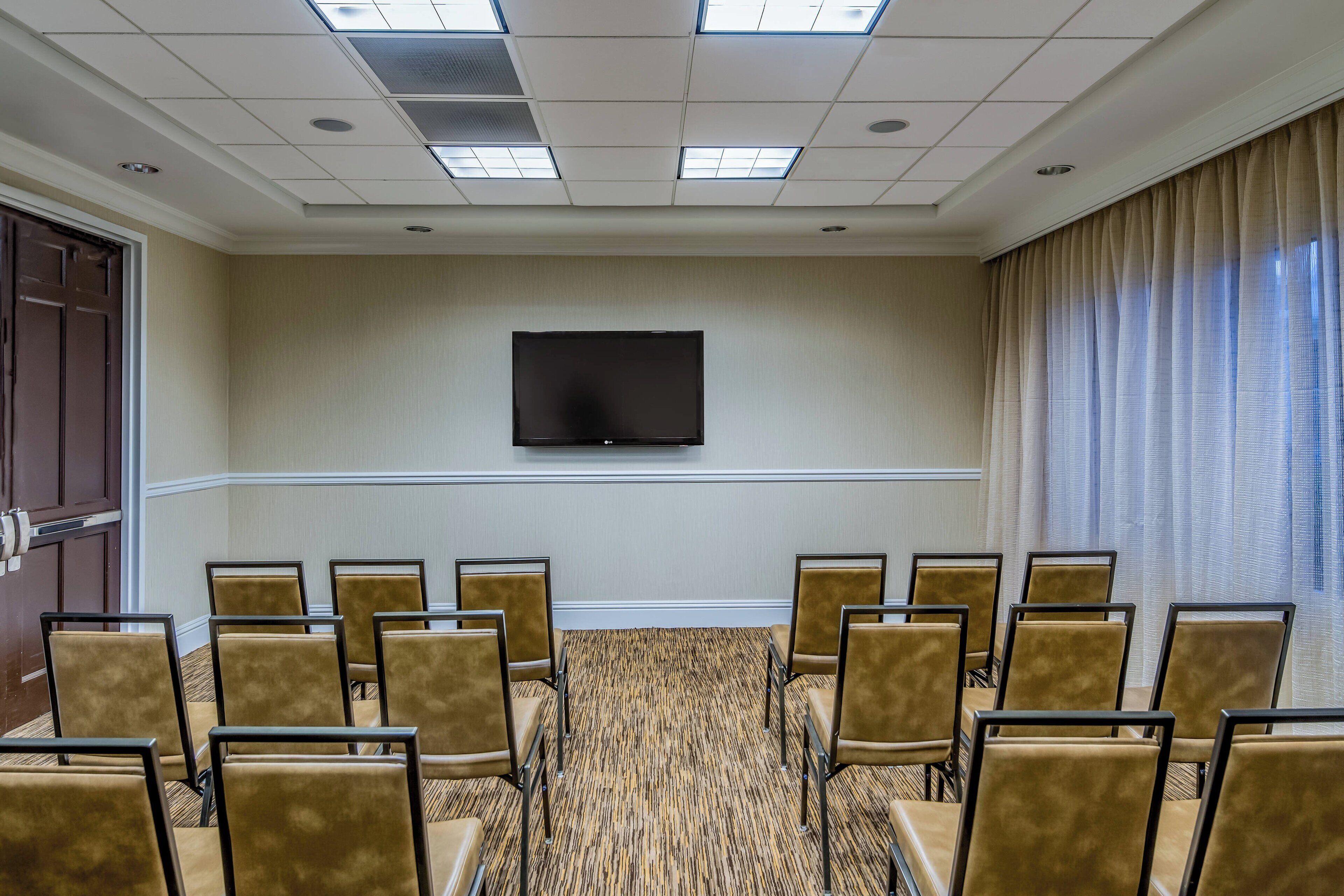 Un salón de conferencias con una televisión de pantalla plana montada en la pared, filas de sillas y piso alfombrado.